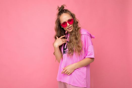 Charming sad young blonde curly woman isolated on pink background wall wearing casual pink t-shirt and stylish colourful sunglasses looking at camera and asking.
