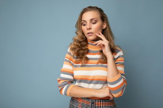 Photo portrait of young pretty beautiful sexy thoughtful blonde woman wearing stylish colourful striped pullover isolated on blue background with copy space and thinking.