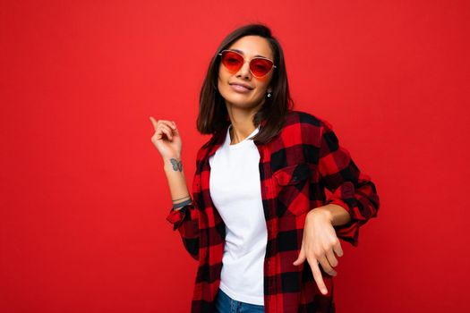 Photo of young beautiful happy smiling brunette woman wearing trendy white t-shirt with empty space for mock up and red check shirt and red sunglasses. Sexy carefree female person posing isolated near red wall in studio with free space. Positive model with natural makeup.