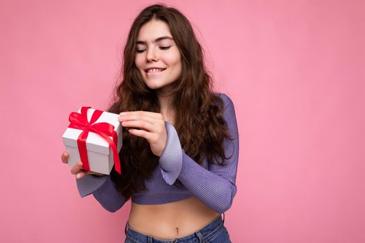 Shot of charming positive smiling brunette curly young woman isolated over pink background wall wearing purple blouse holding white gift box with red ribbon and unboxing present.