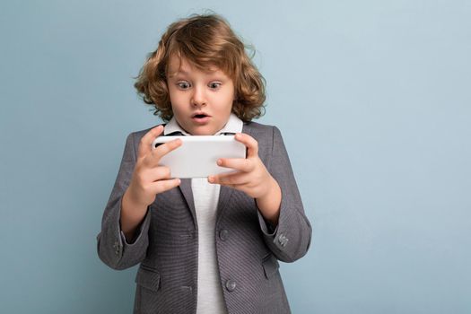 Photo shot of Handsome amazed emotional child boy with curly hair wearing grey suit holding and using phone isolated over blue background looking at smartphone playing games.