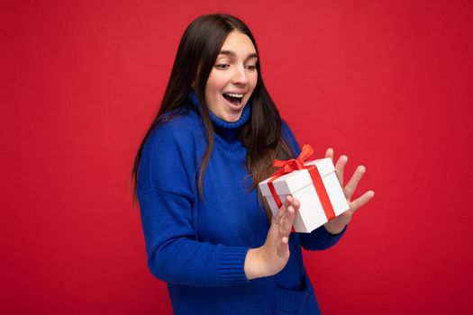 Photo shot of pretty positive surprised young brunet woman isolated over colourful background wall wearing trendy outfit look holding gift box and looking at present box with red ribbon.