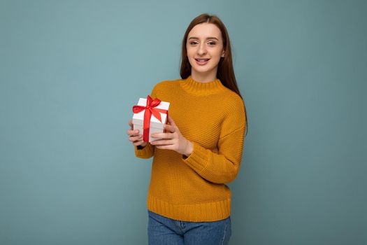 Beautiful happy young brunette woman isolated over colourful background wall wearing stylish casual clothes holding gift box and looking at camera.