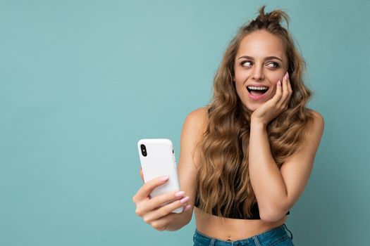 Photo of attractive crazy amazed surprised young woman wearing casual stylish clothes standing isolated over background with copy space holding and using mobile phone looking to the side.