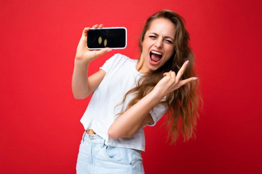 joyful smiling young blonde woman good looking wearing white t-shirt standing isolated on red background with copy space holding phone showing smartphone in hand with empty screen display for mockup looking at camera and showing peace gesture.