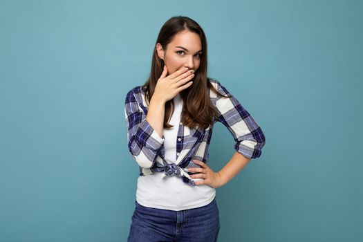 Young positive happy attractive pretty brunet woman with sincere emotions wearing stylish check shirt standing isolated on blue background with empty space and laughing covering mouth.