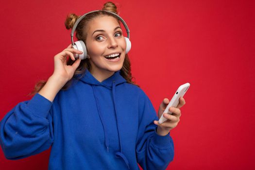 Photo of beautiful overjoyed young woman wearing stylish blue hoodie isolated over red background wearing white bluetooth wireless headphones and listening to music and using mobile phone surfing on the internet looking at camera. empty space