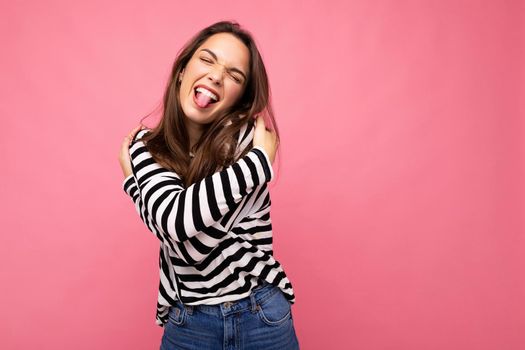 Photo shot of cute attractive pretty young brunette woman wearing casual striped longsleeve isolated over colorful background with copy space having fun and showing tongue. Positive concept