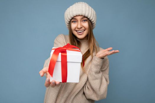 Beautiful happy young dark blonde woman isolated over colourful background wall wearing stylish casual clothes holding gift box and looking at camera.