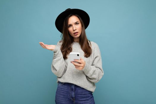 Beautiful young asking dissatisfied brunette woman wearing black hat and grey sweater holding smartphone looking at camera isolated on background.Copy space