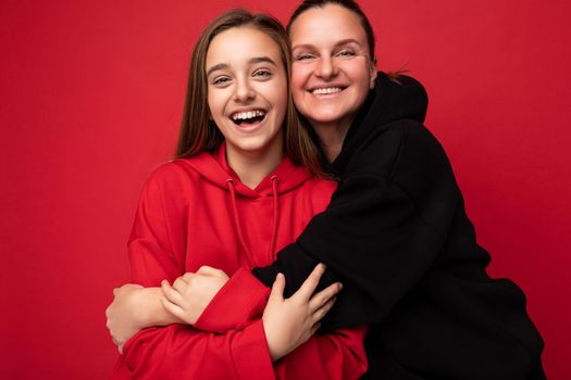 Photo shot of attractive positive smiling brunette female teenager wearing stylish red hoodie and adult brunette woman wearing trendy black hoodie standing isolated over red background wall looking at camera and having fun.