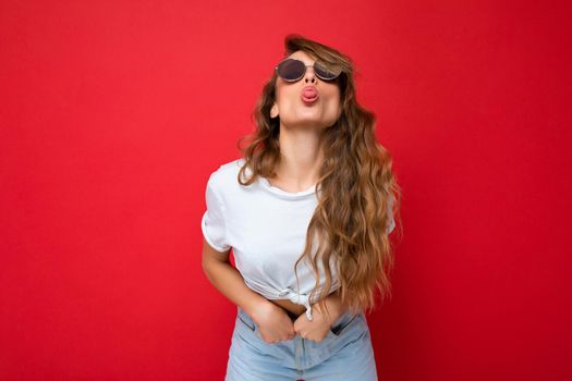 Shot of beautiful sexy young dark blonde curly woman isolated over red background wall wearing casual white t-shirt and stylish sunglasses and giving kiss at camera