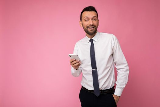 Photo shot of handsome positive good looking young man wearing casual stylish outfit poising isolated on background with empty space holding in hand and using mobile phone messaging sms looking at camera.