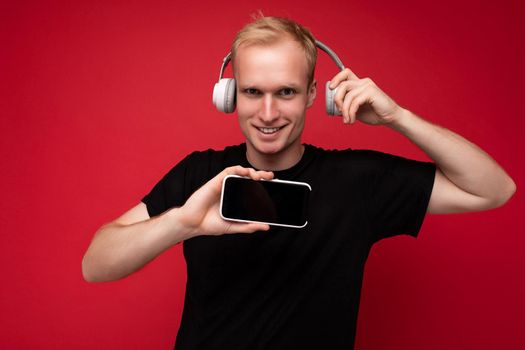 Smiling happy handsome blonde young guy wearing black t-shirt and white headphones standing isolated over red background with copy space holding smartphone and showing mobile phone screen listening to music and having fun looking at camera.