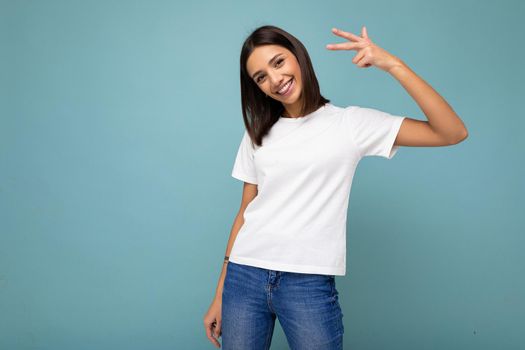 Photo of positive smiling joyful young beautiful brunette woman with sincere emotions wearing casual white t-shirt for mockup isolated over blue background with copy space.