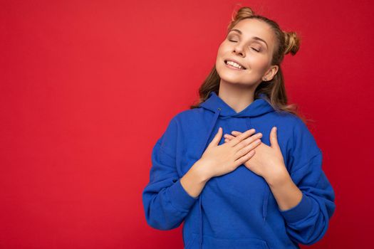 Photo of young positive happy smiling beautiful woman with sincere emotions wearing stylish clothes isolated over background with copy space and holding hands on heart.