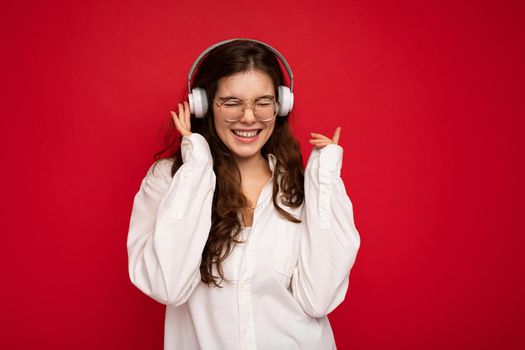 Photo of beautiful happy smiling young brunette woman wearing white shirt and optical glasses isolated over red background wall wearing white wireless bluetooth headphones listening to cool music enjoying.