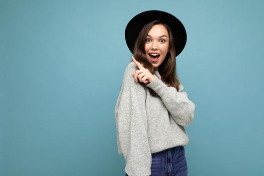 Photo of attractive brunette happy joyful young woman pointing finger at free space for text wearing grey jersey and black hat isolated on blue background.