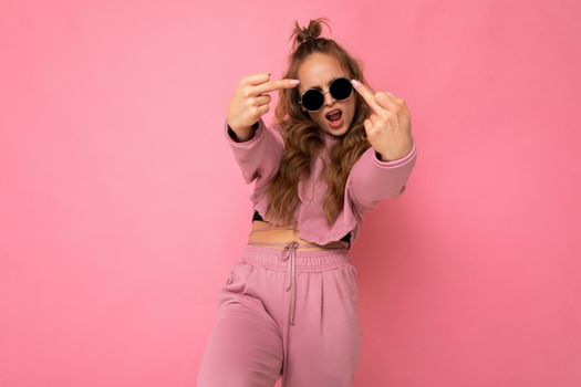 Young happy positive cool beautiful blonde wavy-haired woman with sincere emotions wearing stylish pink sport suit and sunglasses isolated on pink background with copy space and showing fuck.