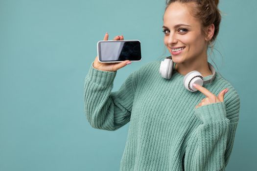 Closeup portrait of attractive positive smiling young woman wearing stylish casual outfit isolated on colourful background wall holding and showing mobile phone with empty screen for cutout wearing white bluetooth headphones and having fun looking at camera and pointing finger at device.