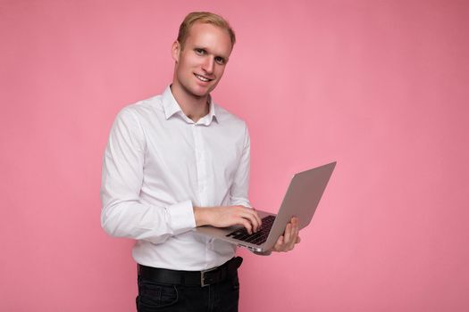 Side profile photo shot of handsome smiling happy confident blonde man holding and using computer laptop typing on keyboard wearing white shirt looking at camera isolated over pink background.