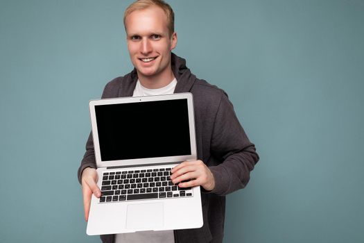 Close-up portrait of handsome smiling blonde man holding computer laptopwith empty monitor screen with mock up and copy space looking at camera isolated over blue background.