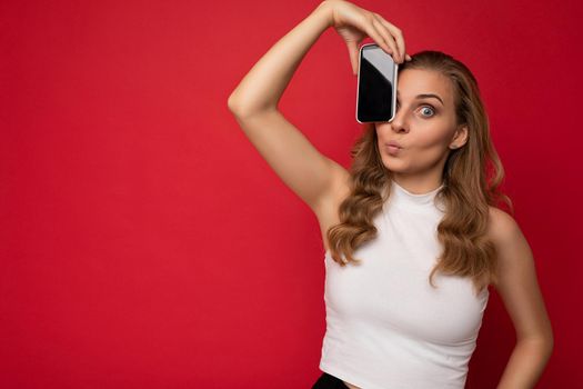 Photo of attractive young blonde female person wearing white t-shirt isolated on red background with copy space holding smartphone showing phone in hand with empty screen for cutout looking at camera.