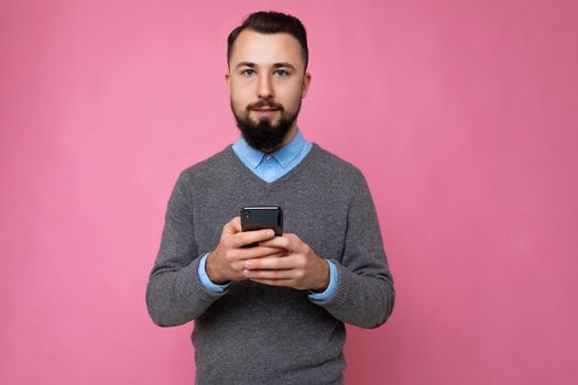 handsome good looking brunet bearded young man wearing grey sweater and blue shirt isolated on pink background with empty space holding in hand and using mobile phone communicating online looking at camera.