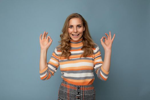Portrait of young attractive blonde curly woman with sincere emotions wearing trendy striped pullover isolated on blue background with copy space and showing ok gesture. It's fine.