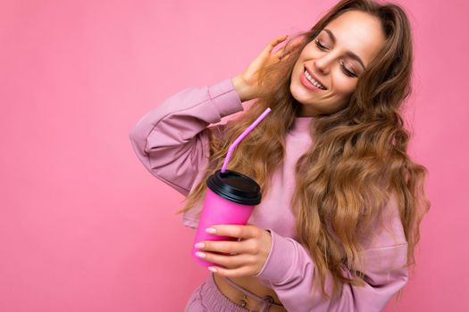 Photo shot of beautiful young smiling blonde woman wearing pink sport clothes isolated over pink background holding paper coffee cup for mockup drinking and enjoying with close eyes.