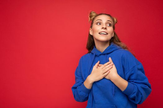 Portrait of young happy positive pretty cute blonde woman with two horns with sincere emotions wearing casual bright blue hoodie isolated on red background with free space and holding hands on chest.