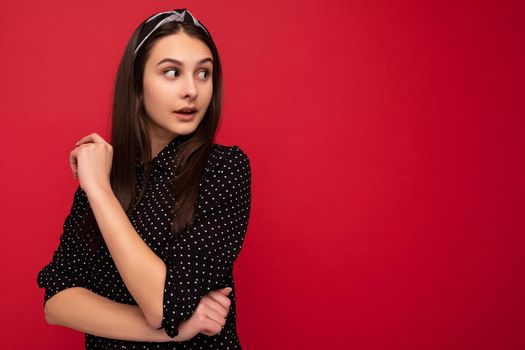 Photo of beautiful surprised positive brunette girl standing isolated over red wall wearing casual stylish black clothes looking to the side.