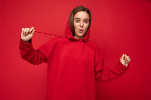 Photo shot of beautiful surprised happy smiling brunette female teenager wearing trendy red hoodie standing isolated over red background wall looking at camera.