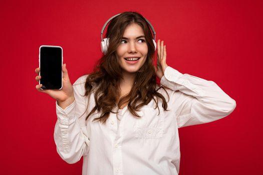 Attractive positive smiling young woman wearing stylish casual outfit isolated on colourful background wall holding and showing mobile phone with empty screen for cutout wearing white bluetooth headphones and having fun looking to the side.