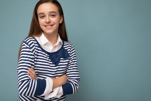 Shot of beautiful positive smiling brunette little girl wearing striped longsleeve standing isolated over blue background wall crossing hands and looking at camera. Empty space