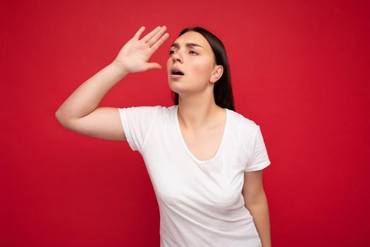 Portrait of emotional young beautiful brunette woman with sincere emotions wearing white t-shirt for mockup isolated over red background with free space and looking to the side with hand.