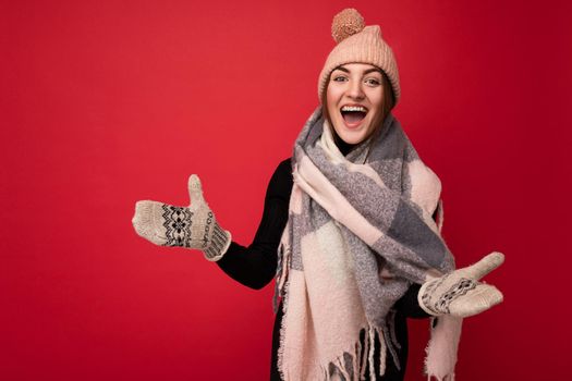 Photo of beautiful happy amazing funny young brunette woman isolated over red background wall wearing winter scarf mittens and warm hat looking at camera and having fun.