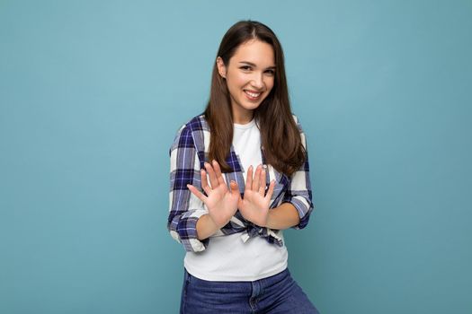 Young positive smiling beautiful brunette woman with sincere emotions wearing trendy check shirt standing isolated on blue background with empty space and showing stop gesture saying no.