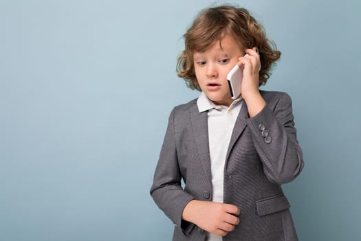 Photo of Handsome boy with curly hair wearing grey suit holding and using phone isolated over blue background looking downand talking on mobile with somebody. Copy space