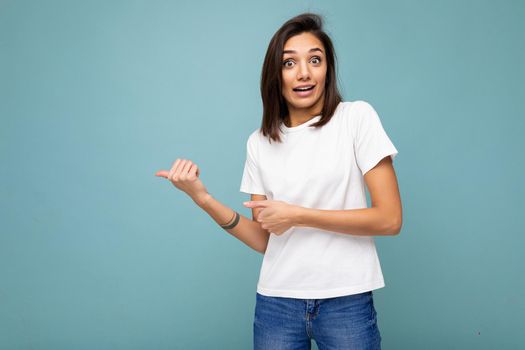 Portrait of shocked amazed young attractive brunette woman with sincere emotions wearing white t-shirt for mockup isolated over blue background with copy space and pointing at free space for text.