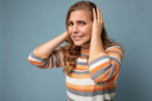 Photo of young emotional angry pretty nice blonde curly woman with sincere emotions wearing casual striped pullover isolated on blue background with free space and covering ears trying not to hear.