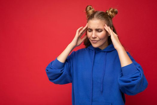 Photo of tired weary young nice pretty blond woman with two horns with sincere emotions wearing casual bright blue hoodie isolated over red background with copy space and having headache. Pain concept.