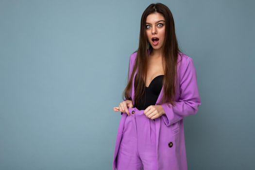 Portrait of shocked amazed beautiful fashionable brunette woman in casual trendy violet jacket isolated on blue background with free space. Surprise concept.