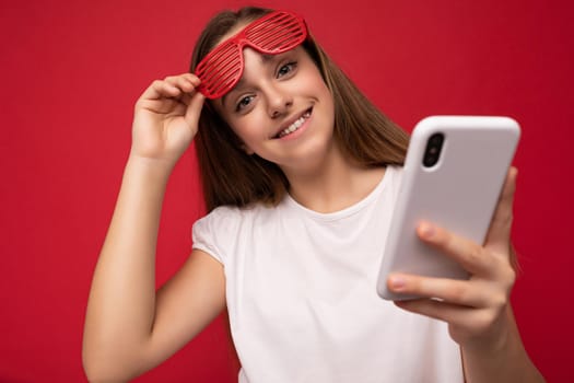 Charming positive young girl wearing white t-shirt and red glasses poising isolated on red background with empty space holding in hand and using mobile phone communicating online looking at camera.