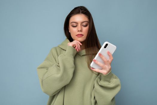 Photo of serious thoughtful Beautiful young brunette woman wearing stylish green hoodie using mobile phone writing message isolated on background looking at smartphone display.