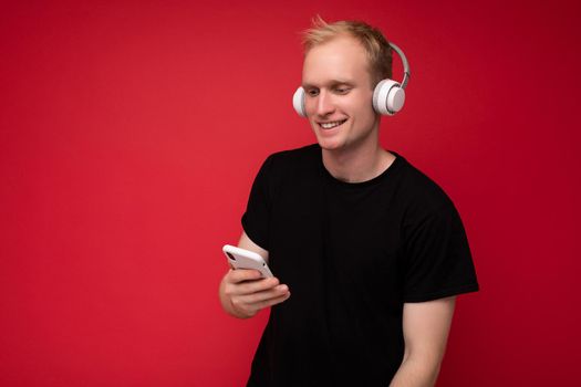 Photo shot of handsome positive good looking young man wearing casual stylish outfit poising isolated on background with empty space holding in hand and using mobile phone messaging sms looking at smartphone display screen.