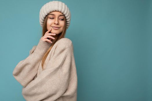 Shot of sexy pretty happy young dark blonde woman standing isolated on blue background wall wearing beige warm sweater and winter beige hat looking down. Free space, copy space