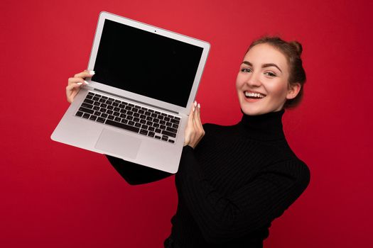 Close-up portrait of Beautiful smiling happy young woman with gathered hair holding computer laptop with empty monitor screen with mock up wearing black longsleeve looking at camera.