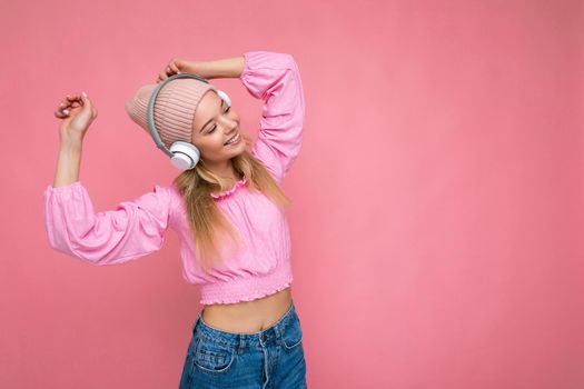 Beautiful happy smiling young blonde woman wearing pink blouse and pink hat isolated over pink background wall wearing white wireless bluetooth headphones listening to cool music having fun and dancing. copy space