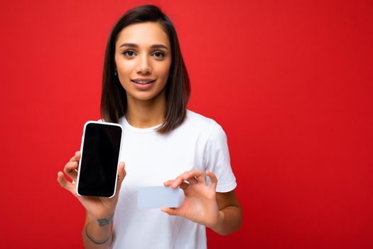 Portrait photo of brunette young woman wearing everyday stylish white t-shirt isolated on red background wall holding and using phone with empty screen for mockup and credit card making payment looking at camera.